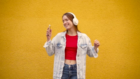 pretty young woman listening to music and dancing in the street with a yellow wall behind