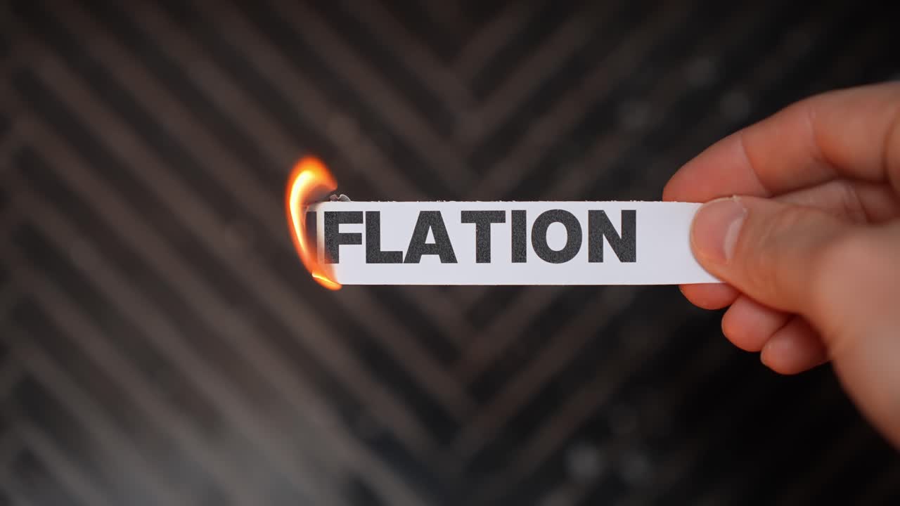 Hand burns a paper label reading “INFLATION,” symbolizing inflation ending—price stability, affordability, and relief for household budgets. Dark background with copy space
