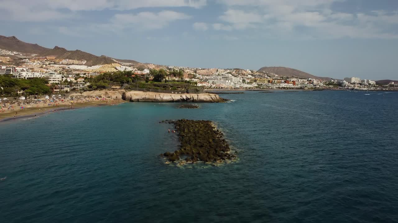 impresionante toma de drones del mar azul en la playa al sur de tenerife los cristianos en el agua de la playa golpeando las rocas con un bonito paisaje con montañas en la parte de atrás