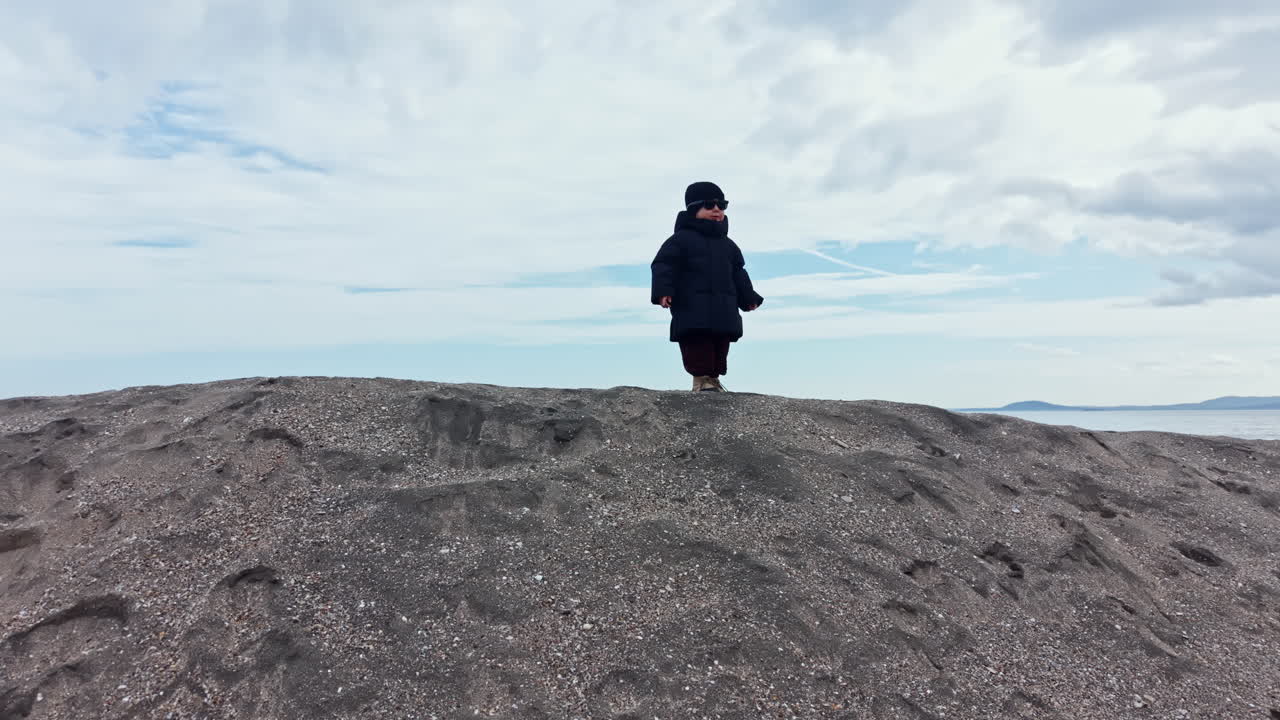 Bambino di 2 anni con giacca e cappello invernali cammina su una spiaggia sabbiosa. Il bambino gioca in riva al mare in inverno. Abbigliamento invernale per bambini. Passeggiata invernale di un bambino al mare