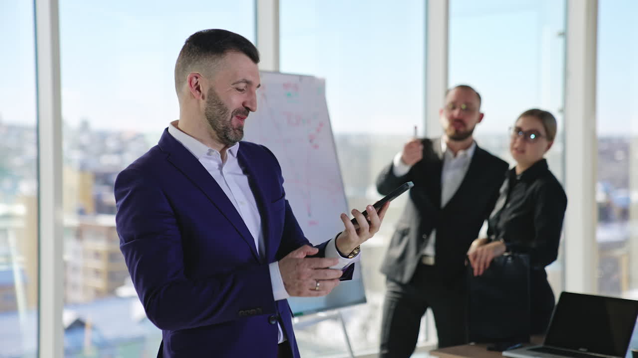 Joyful positive man in suit talking on the phone and smiling. Colleagues behind address him saying something funny, all starting to laugh. Blurred backdrop.