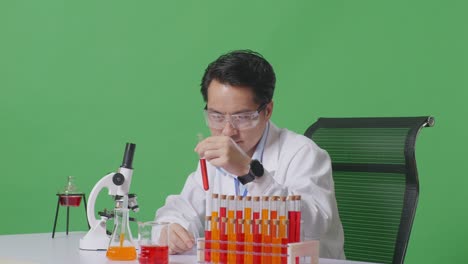 side view of asian man scientist looking at test tube then smiling and showing thumbs up gesture while working on the table with microscope in the green screen background laboratory