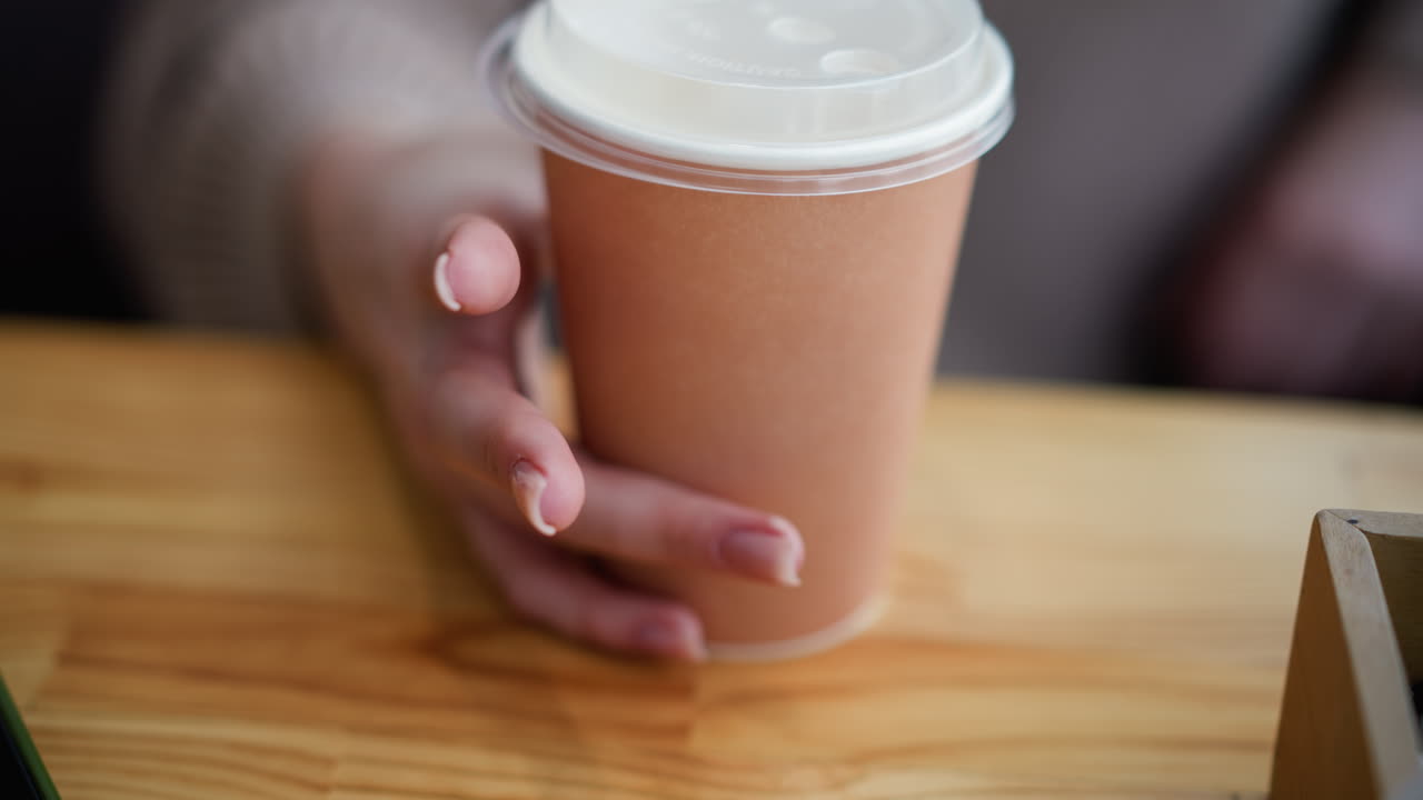 vista de cerca de la mano de la dama tocando la taza de café en la mesa de madera con el teléfono parcialmente visible y la caja de madera en el fondo, creando una atmósfera acogedora con un borrón suave