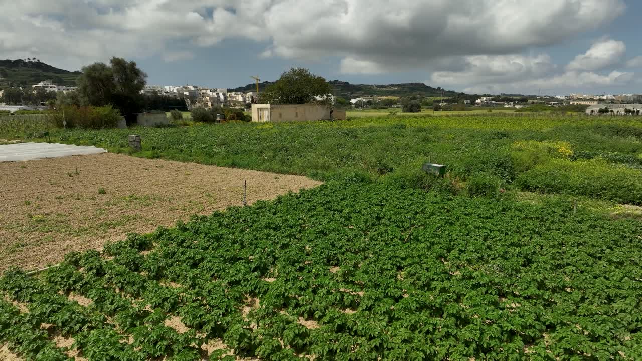 Drone glides low above lush green fields and a rustic stone shed in Malta, with distant villages and rolling hills beneath a dramatic sky, highlighting the island’s rural landscape.