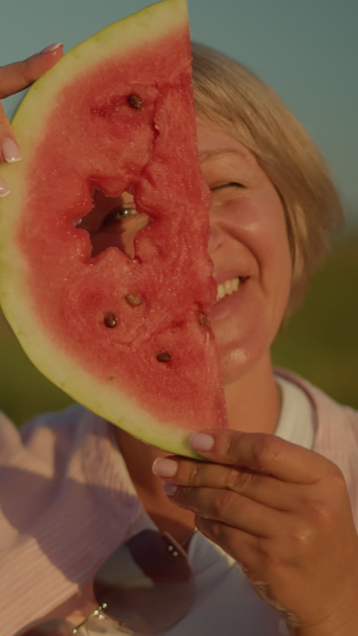 mujer sosteniendo una rebanada de sandía con un recorte en forma de estrella mientras sonríe juguetón, mirando a través del recorte bajo la luz del sol brillante, el fondo presenta un paisaje verde borroso