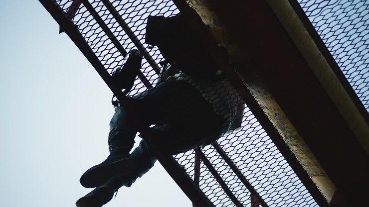 Uomo bianco appeso a una grata di metallo, esploratore urbano appollaiato su una passerella arrugginita, gambe che penzolano sopra la volta di un albero, cielo retroilluminato, texture grintosa, alta tensione, silhouette ad angolo basso, atmosfera avventurosa