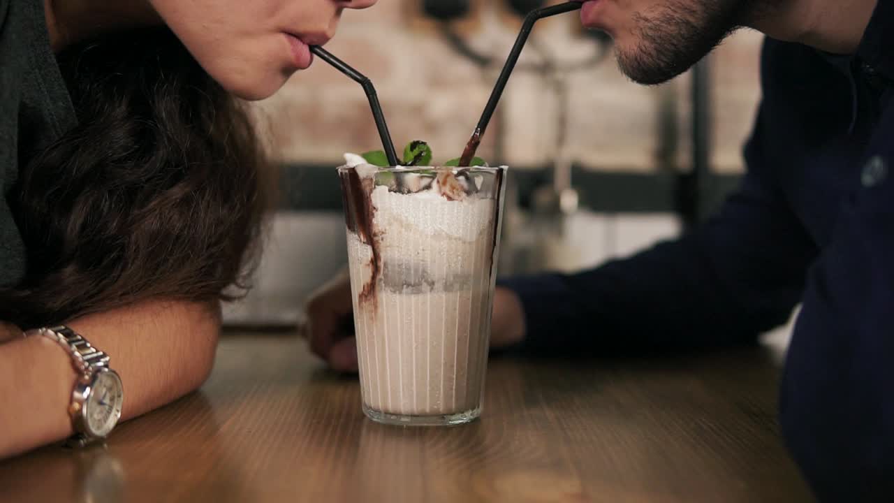 Close Up view of attractive young couple in cafe sitting at the wooden table and sharing milkshake drinking it together using two