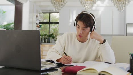 asian male teenager with headphones learning and using laptop in living room