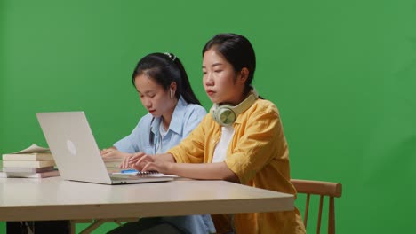 asian woman student is bored while sitting with her friend and typing on a laptop on the table in the green screen background classroom