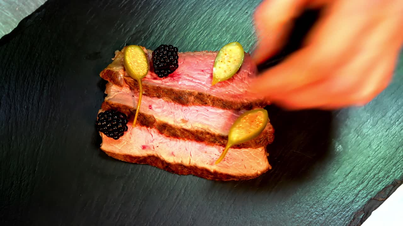 Primo piano delle mani che impiattano con cura un piatto gourmet con carne affettata guarnita con bacche e decorazioni su un piatto di ardesia.