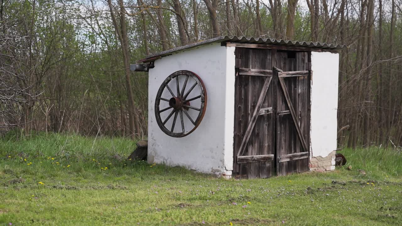 Small white shed with wooden door and wagon wheel in grassy area
