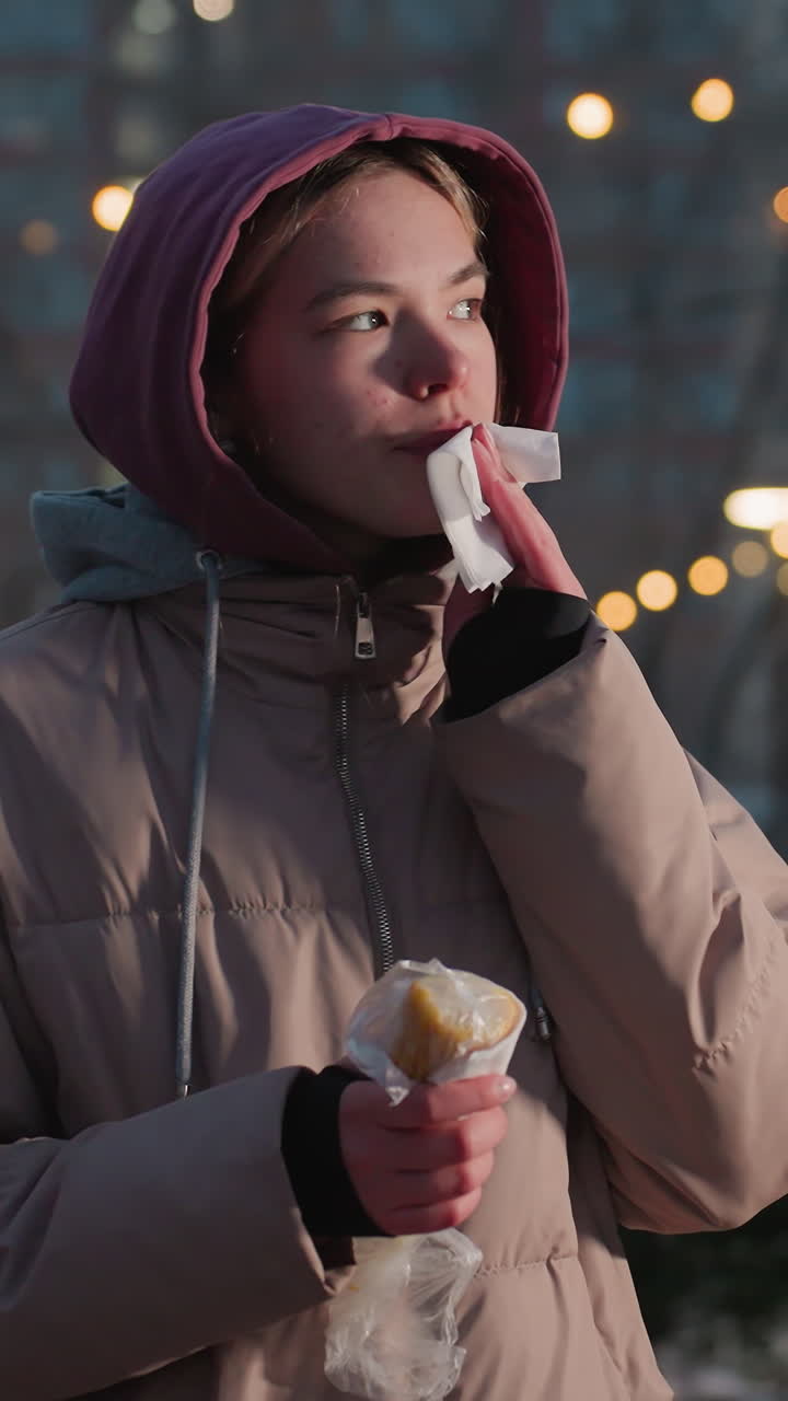 niña joven sosteniendo un bocadillo con una mano mientras usa una servilleta para limpiar la boca en un entorno al aire libre de invierno, luces bokeh suaves en el fondo, disfrutando de la comida en un día frío con personas caminando cerca