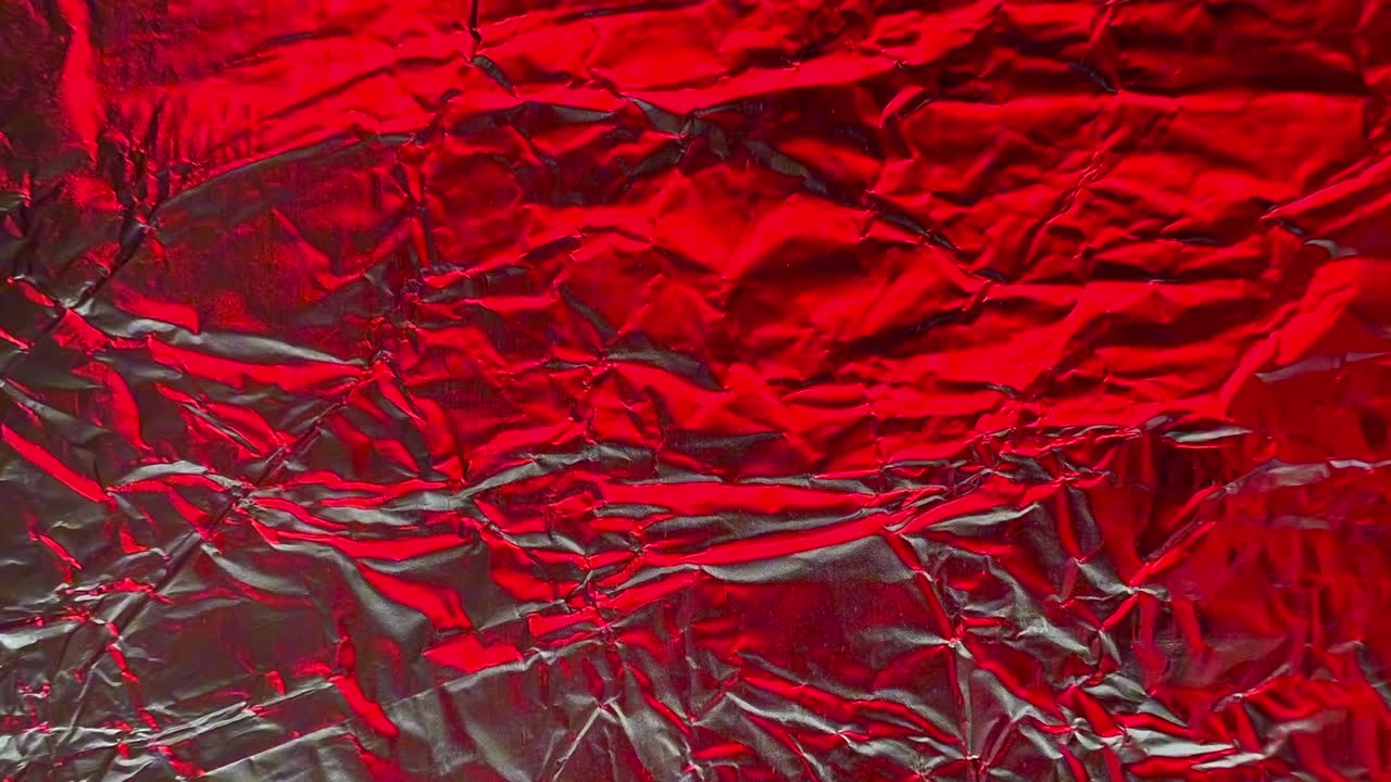 Shiny crumpled red foil with visible wrinkles and folds creates an abstract textured background, ideal for design projects needing a metallic or reflective surface