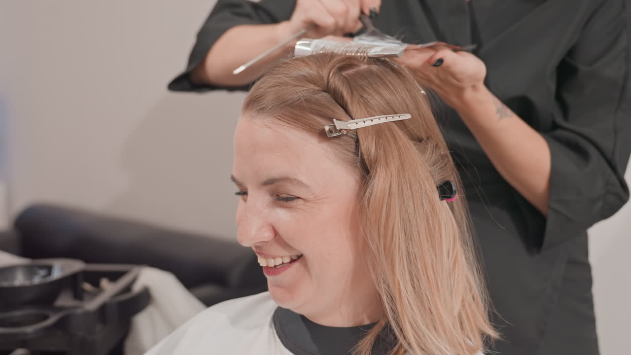 Lächelnde weiße Kundin mit Folien-Strähnchen auf blondem Haar im Salon lacht während einer freundlichen Farbbehandlung mit Stylist. Clips und Kamm sichern die Partien, warmes Studiolicht, Energie bei der Beratung, Colorist