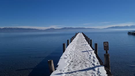 Sunny-Winter-Day-on-Coast-of-Lake-Tahoe-USA,-Snow-Capped-Dock,-Calm-Blue-Water-and-Mountains-in-Horizon