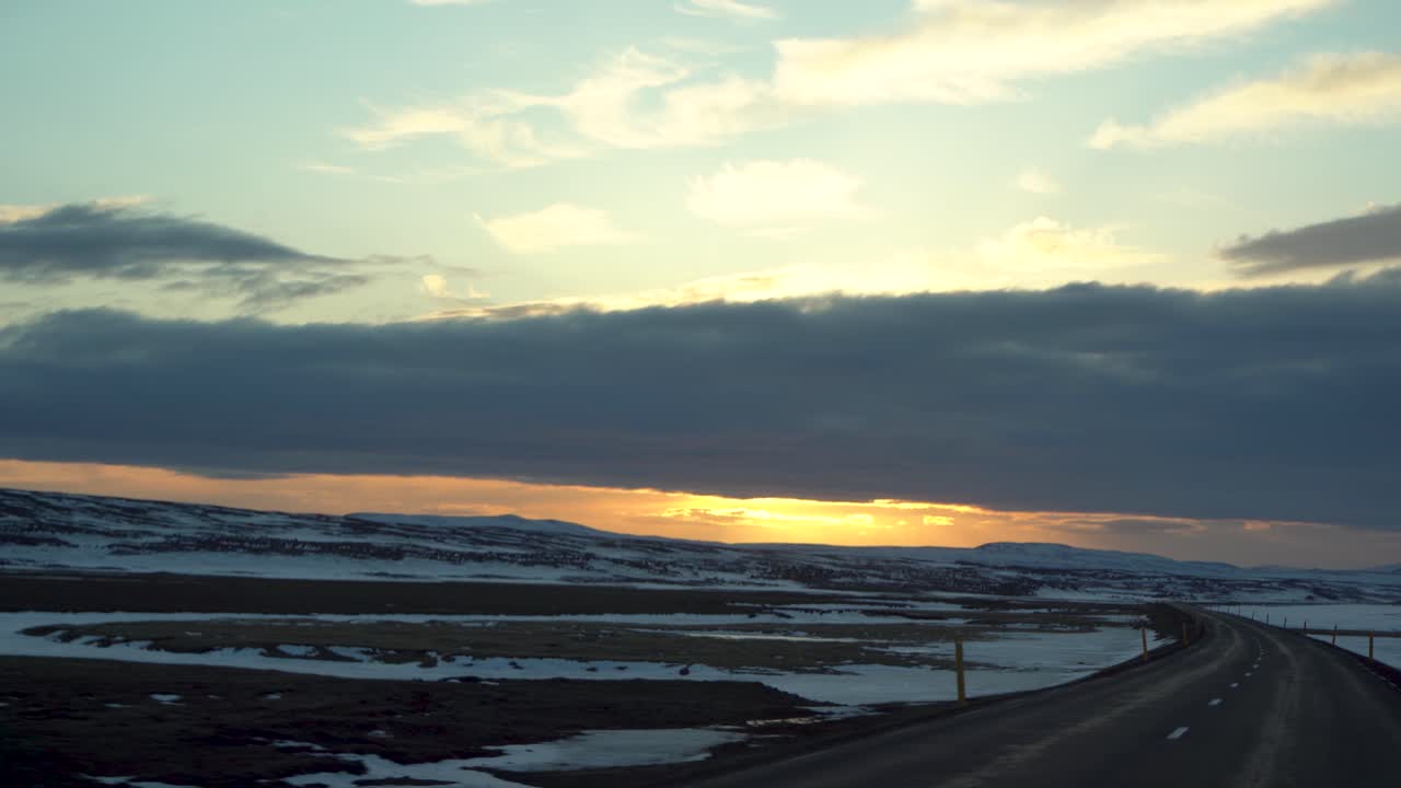 el crepúsculo se asienta sobre un sereno paisaje islandés visto desde un coche de conducción, manchas de nieve salpicando el vasto terreno bajo un cielo dramático