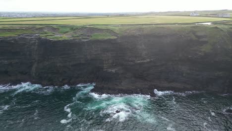 Acantilados-De-Kilkee-Con-Olas-Rompiendo,-Exuberantes-Campos-Verdes-En-El-Fondo-Desde-La-Perspectiva-De-Un-Dron