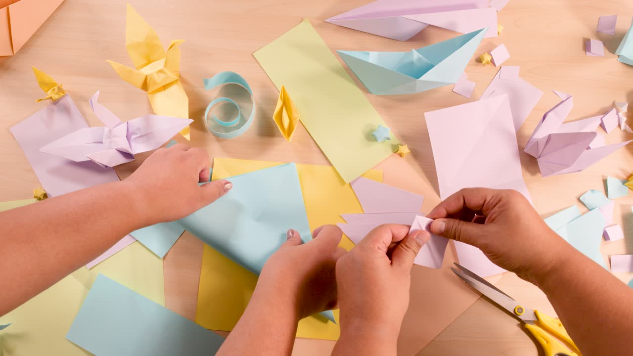 Two people fold pastel paper for origami on a desk, top-down view, soft lighting