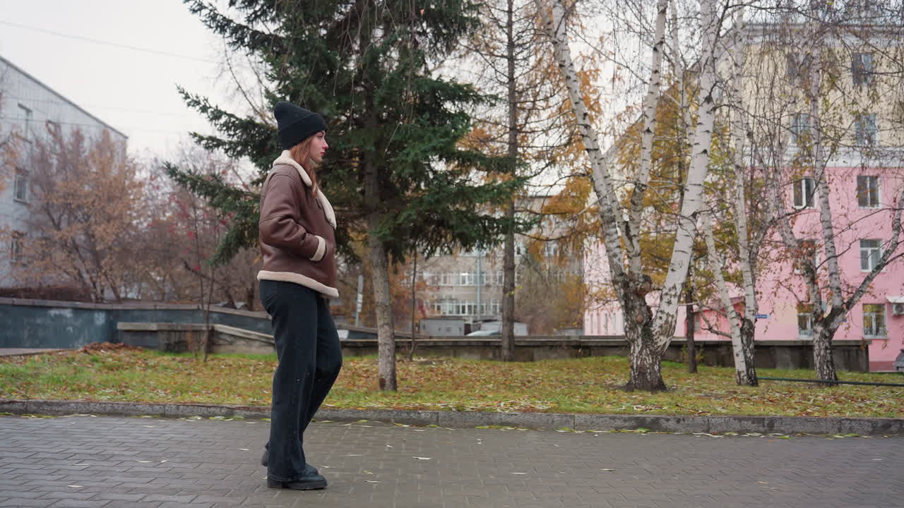 Side view of student in black knit cap, brown shearling jacket, and black trouser walking calmly through paved urban park on cold overcast day with light snowfall and colorful autumn background
