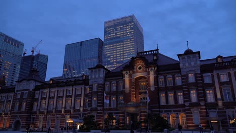 Berühmter-Tokyo-bahnhof-Bei-Nacht-Mit-Pendlern,-Die-Sich-Bewegen,-Und-Wolkenkratzern-Im-Hintergrund---Seitliche-Ansicht-Aus-Niedrigem-Winkel