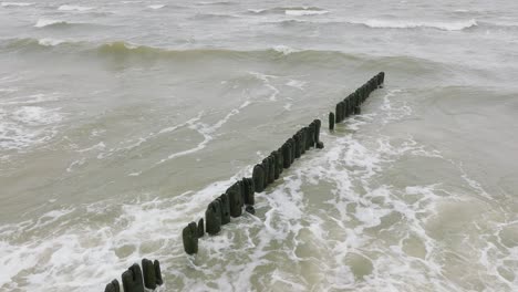 Vista-Aérea-De-Pájaros-De-La-Costa-Del-Mar-Báltico-En-Un-Día-Nublado,-Antiguo-Muelle-De-Madera,-Playa-De-Arena-Blanca,-Grandes-Olas-De-Tormenta-Aplastando-La-Costa,-Cambios-Climáticos,-Tiro-Medio-De-Drones