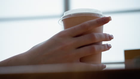 side hand view of woman with well-manicured nails tapping coffee cup on wooden table with blurred background featuring warm lighting, creating a cozy and aesthetic ambiance