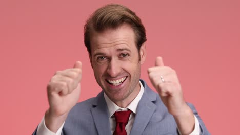 proud young man in elegant suit finding out good news while looking down, feeling joyful and clapping on pink background in studio