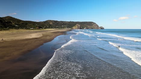 Panorámica-A-Través-De-La-Playa-De-Arena-Negra-De-Piha-Con-El-Reflejo-De-Un-Surfista-Saliendo-Del-Agua-Del-Océano