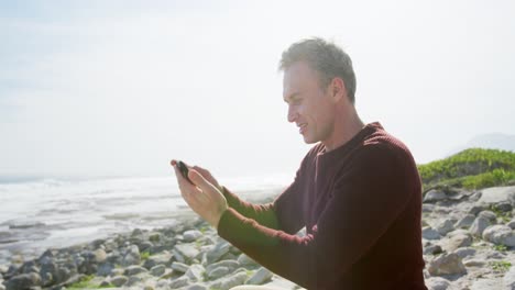 Hombre-Caucásico-Disfrutando-De-Tiempo-Libre-Junto-Al-Mar-En-Un-Día-Soleado-Sentado-En-Un-Teléfono-Inteligente