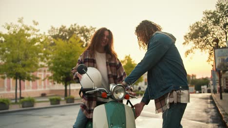 the girl turns on and shirts with curly brown hair gets on her green moped, puts on a white helmet, her boyfriend with curly hair in a denim shirt walks next to her, puts an olive helmet on his head and does not start riding a moped on the street