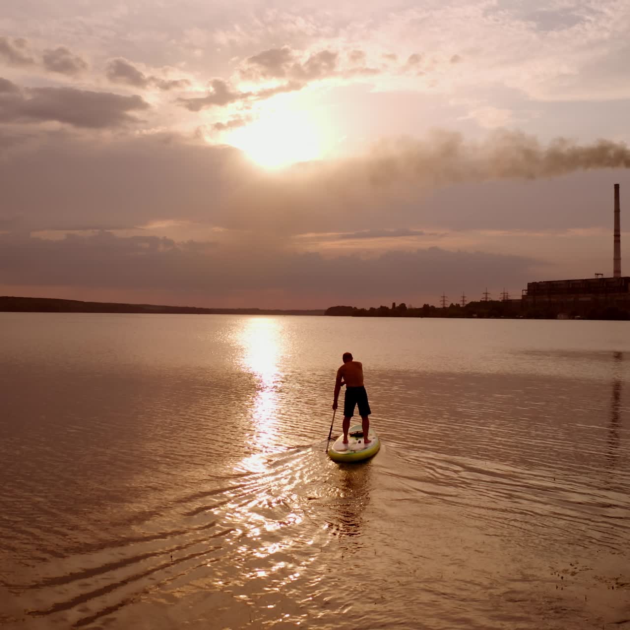 Paddleboarding al tramonto. Ragazzo sportivo che naviga su una tavola con remi sull'acqua serale sullo sfondo di ciminiere industriali con fumo. Viaggio acquatico