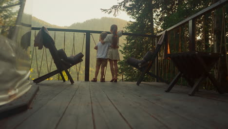 brother and sister watch nature of calm morning forest hugging on balcony of glamping house. boy and girl together rest enjoying deep atmosphere