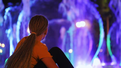 the child admires the fountain with lighting, sits on the edge of the fountain, rear view