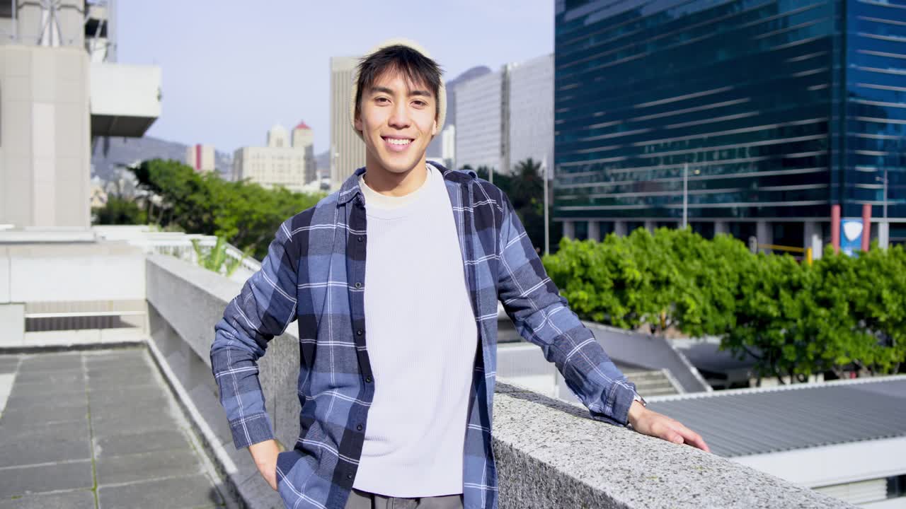 옥상 테라스에서 걷는 아시아 남성, 난간에 기대어 유리 건물과 나무를 둘러보며 미소 짓는 모습.