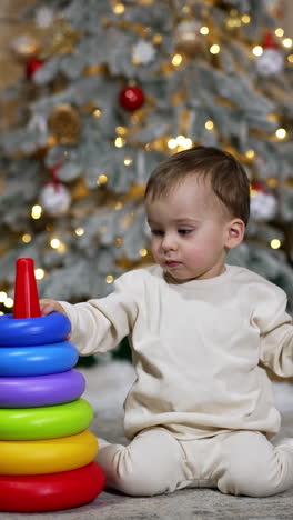 lovely caucasian baby building a toy pyramid. toddler sits on the floor not far from the christmas tree. vertical video