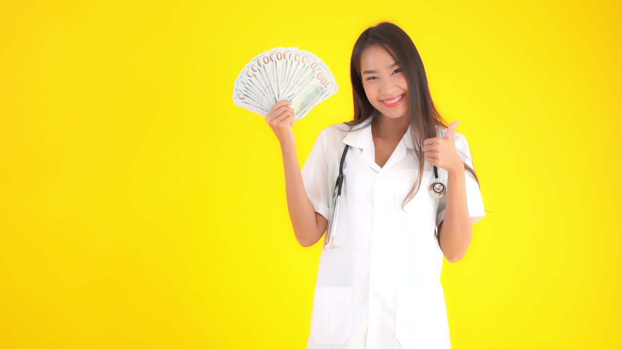 Portrait of asian female doctor showing dollar bills. Huge sallary for medical workers concept, static slow motion, isolated on yellow background