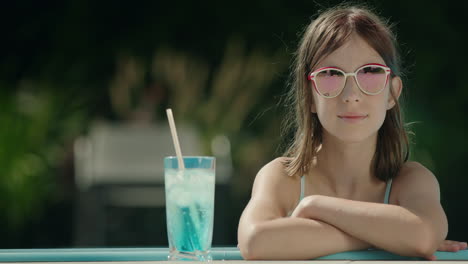 portrait of a cute little girl in sunglasses by the side of the pool. nearby is a cool cocktail