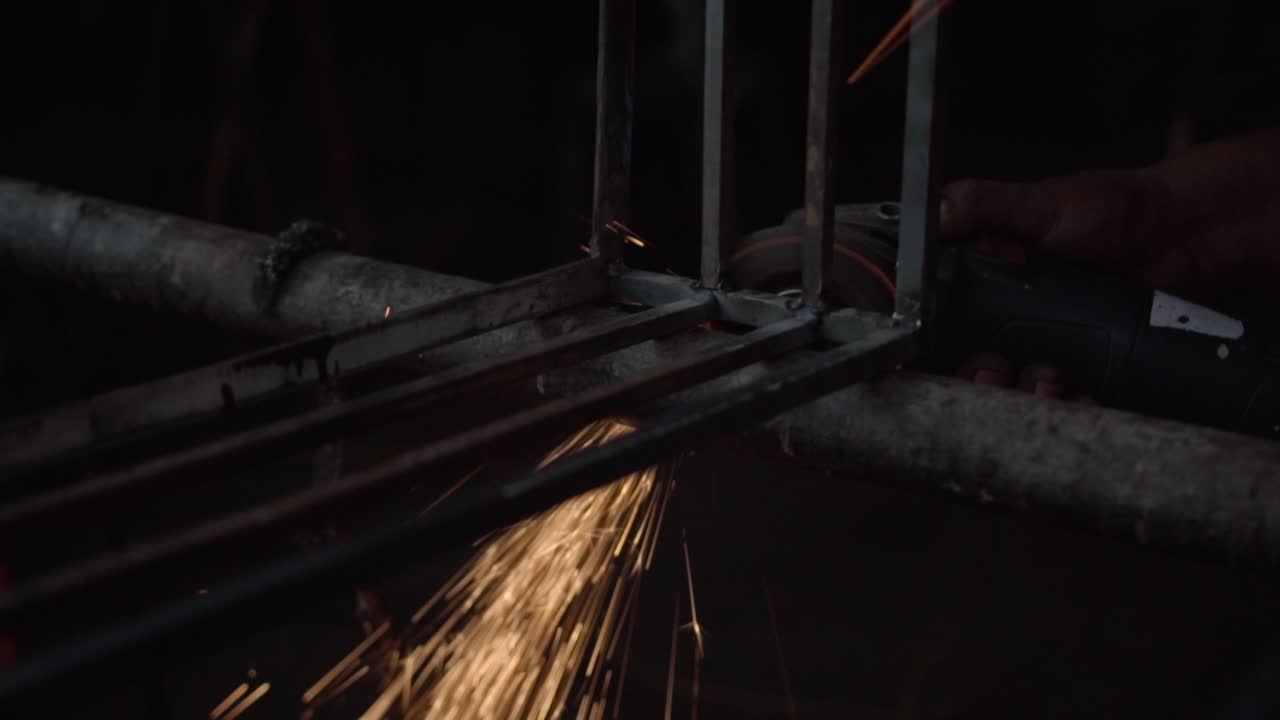 Operaio industriale che taglia barre d'acciaio con una smerigliatrice angolare mentre scintille luminose volano all'interno di un'officina metallurgica