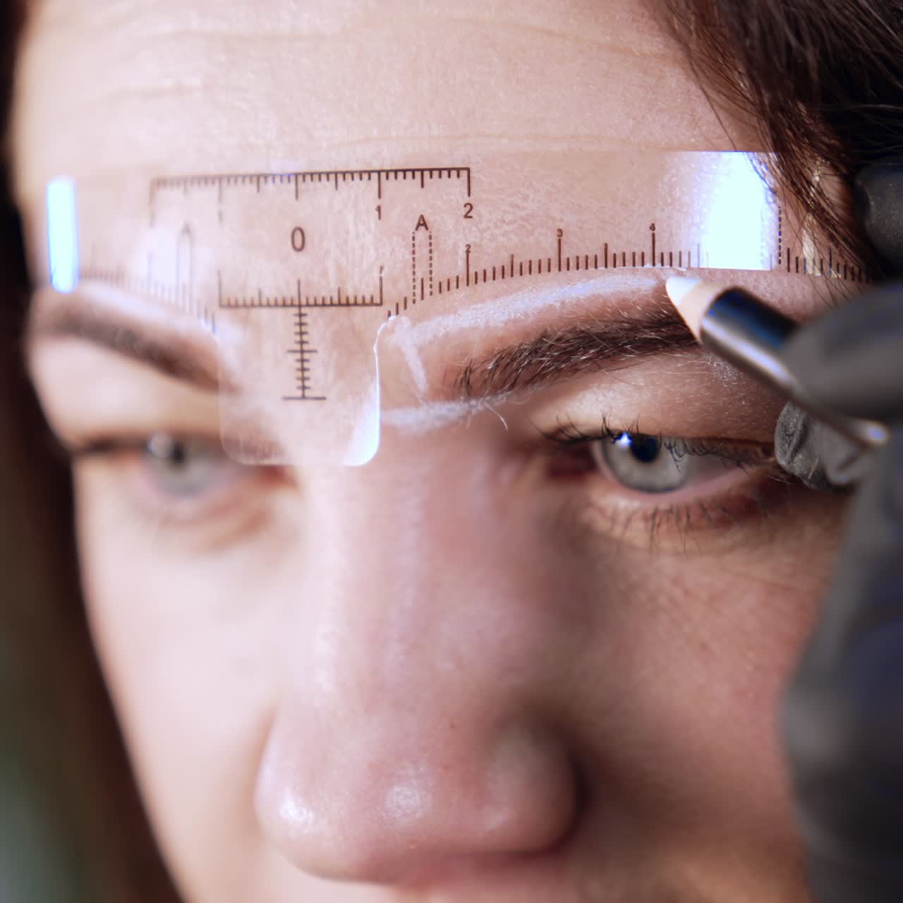 Upper part of an adult brunette female face. A ruler is stuck to the forehead and gloved hand draws white lines for eyebrow permanent make up. Close up