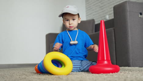 beautiful caucasian toddler sits on the floor waving his hands from side to side. happy boy starts to collect toy pyramid. low angle view.