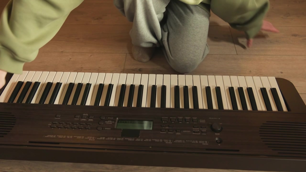 Uppifrån-perspektiv av en person som sitter i skräddarställning bakom ett elektroniskt keyboard, med händerna svävande över svarta och vita tangenter under en avslappnad paus efter att ha spelat, i en lugn och varm köksmiljö.