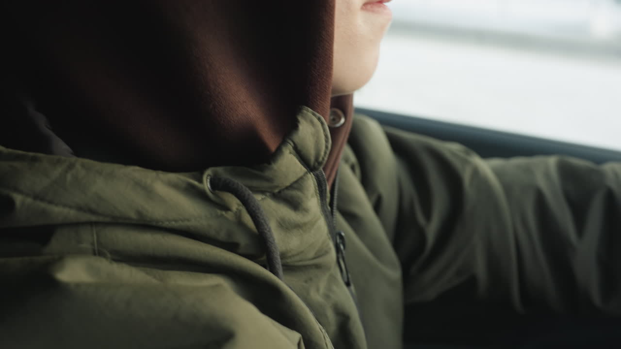close up of student in hoodie and glasses sitting inside car staring quietly outside with pensive expression and blurred snowy background conveying thoughtful mood in cold winter urban environment