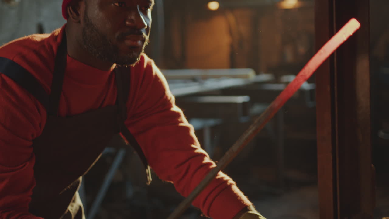 herrero afroamericano calentando la barra de acero con fuego en la forja de carbón