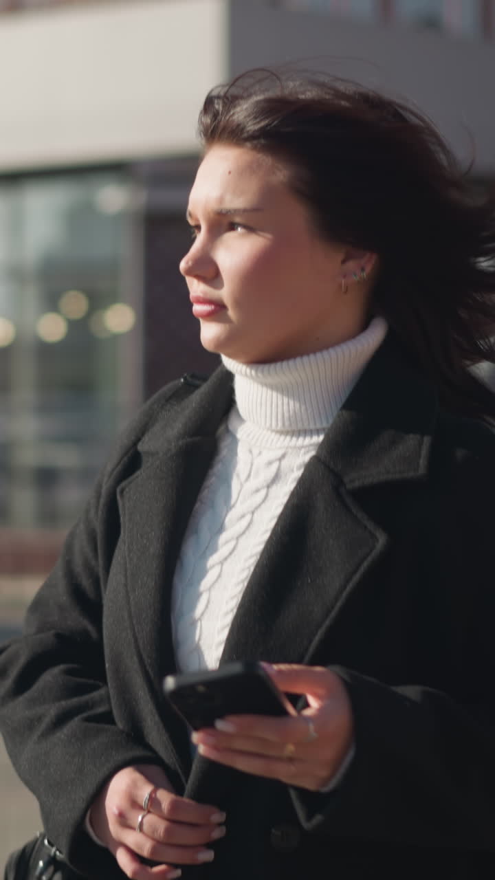 Lady walking outdoors in a black coat and white turtleneck, holding her phone and glancing around with a thoughtful expression, background features modern urban buildings
