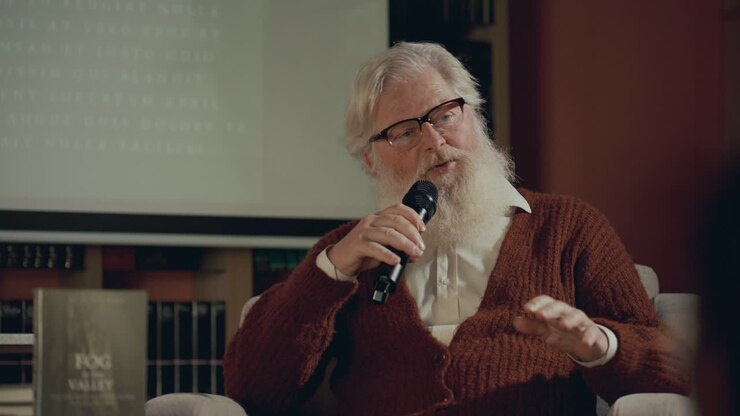autore anziano che parla in microfono dal palco durante una lezione o una discussione su un libro