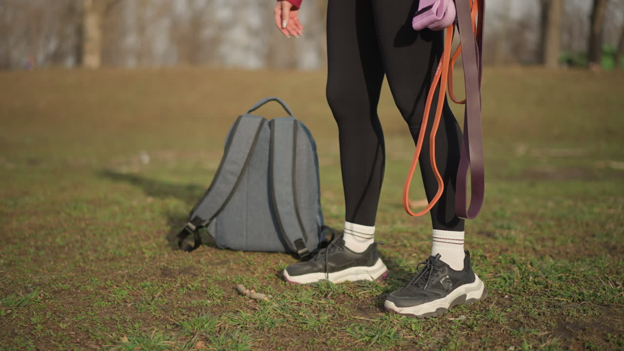 Asian Woman Walking In Park Holding Backpack And Colorful Leashes, Relaxed Pace Across Grassy Slope, Hoodie And Leggings Outfit, Soft Light And Natural Outdoor Calm Emphasize Leisure And Routine Care