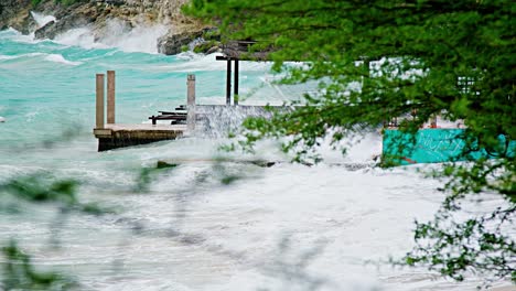 Zoom-shot-with-plants-in-foreground-of-local-wooden-fishing-boat-being-smashed-into-land-during-sudden-storm-with-rough-waves,-Caribbean