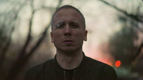 a man in a green jacket smoking a cigarette outdoors at dusk, the background is blurred with a soft, warm light