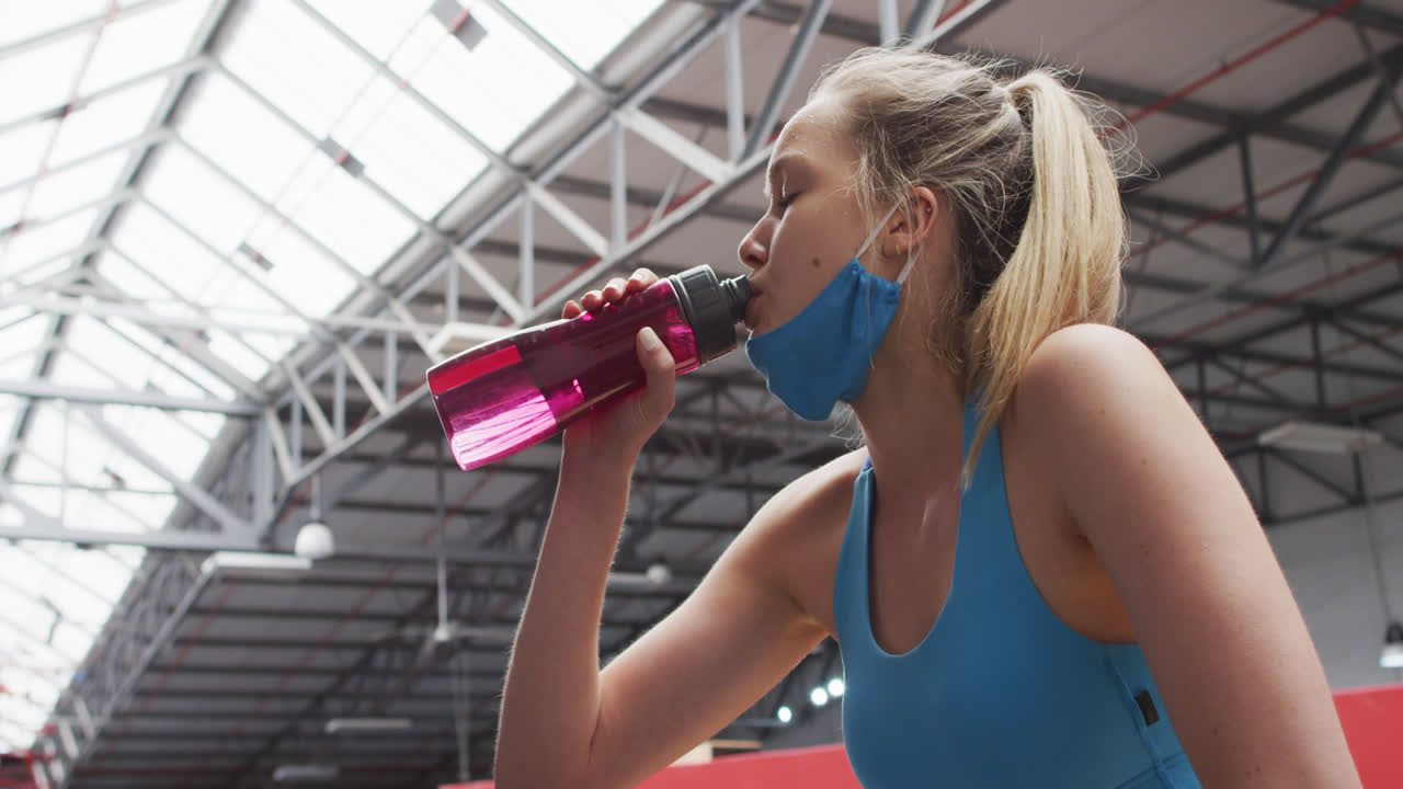mujer caucásica con máscara de cara baja bebiendo agua en el gimnasio