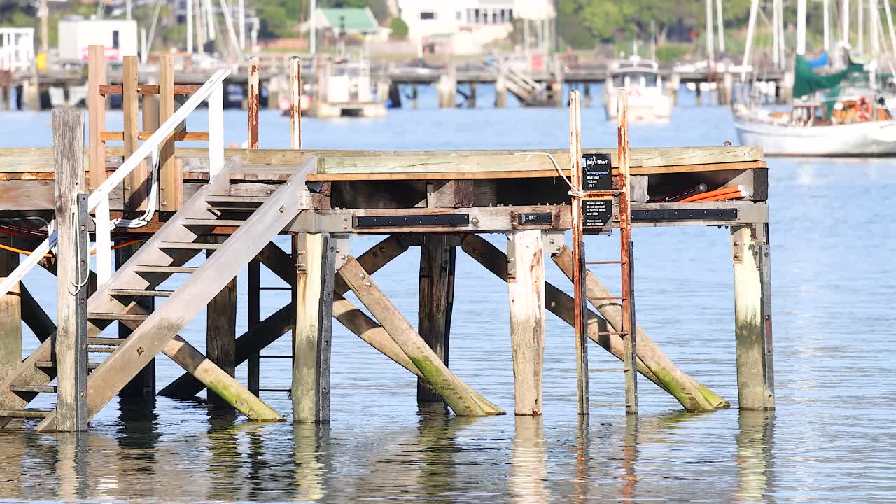 Wooden pier in Akaroa, New Zealand, with calm waters and distant boats. Bright daylight enhances the serene coastal atmosphere